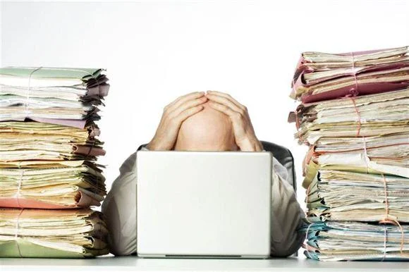 man holding his head in front of a computer surrounded by stacks of paper